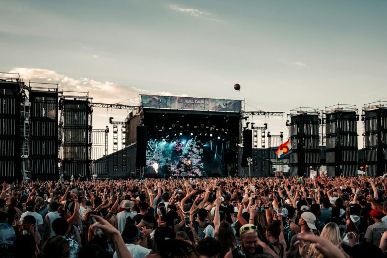Large outdoor music festival crowd facing a brightly lit stage with rigging, banners, and raised hands at sunset/ dusk.