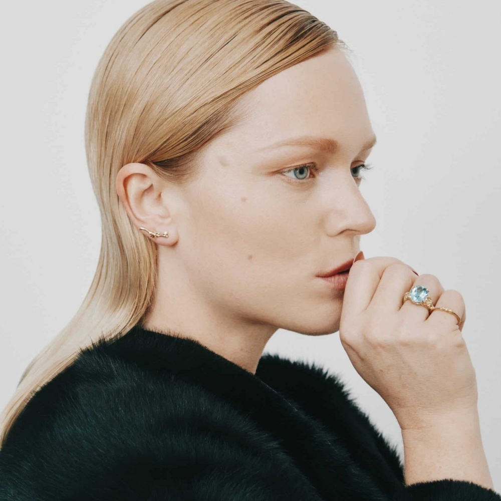 Side profile of a blonde woman wearing a black fur stole, touching her lips with her fingers and a blue gemstone ring on her finger.