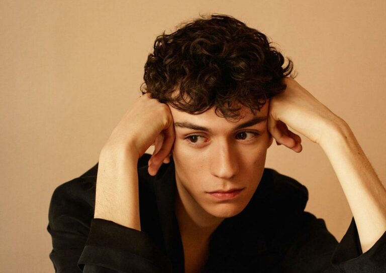 Young man with curly hair rests his head in his hands, gazing pensively to the side against a beige backdrop.