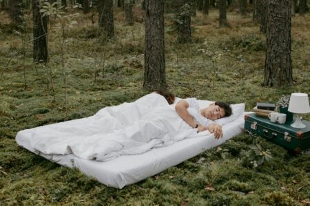 Person sleeping on a white mattress in a forest, with a green vintage suitcase, lamp, books, and mugs nearby.
