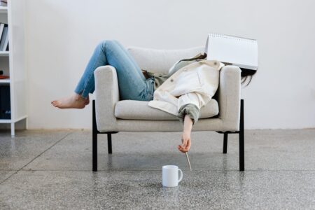 Person lounging on a light sofa with a notebook covering their face, one arm reaching toward a white mug on the floor, pencil in hand.