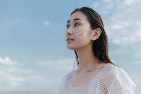 Woman with wet hair wearing a white mesh top, looking up at the sky, with skincare cream on her cheek and a tattoo on her collarbone, by the beach.
