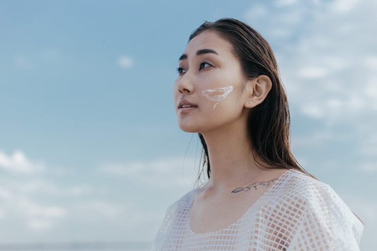 Woman with wet hair wearing a white mesh top, looking up at the sky, with skincare cream on her cheek and a tattoo on her collarbone, by the beach.
