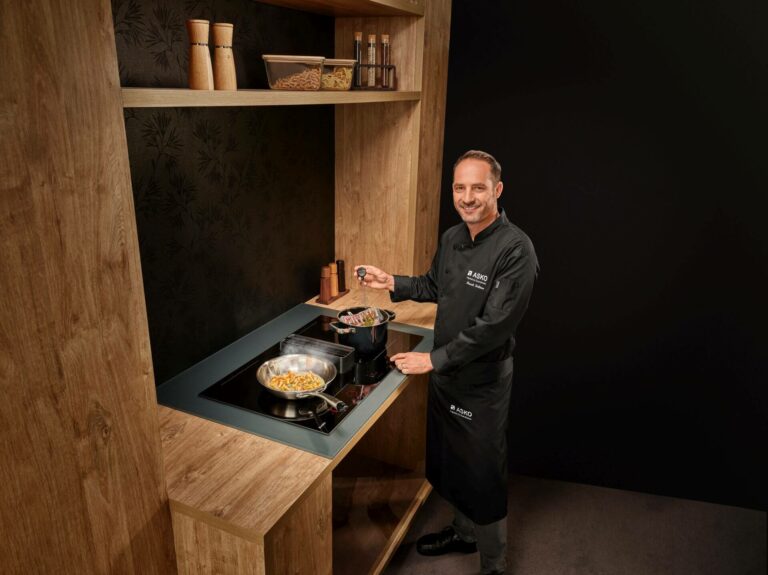 Male chef in a black uniform cooks at a wood-framed kitchen station, using an induction cooktop with pasta in a pan on the hob.