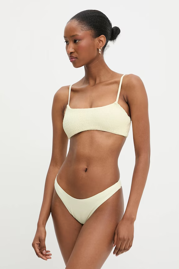 Woman modeling a cream-colored bikini with a textured bandeau-style top and matching bottoms, standing against a white studio backdrop.