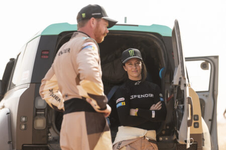 Race crew members stand at the open door of a large off-road vehicle; a woman wearing a Monster cap sits inside, while a man in beige racing gear stands nearby.