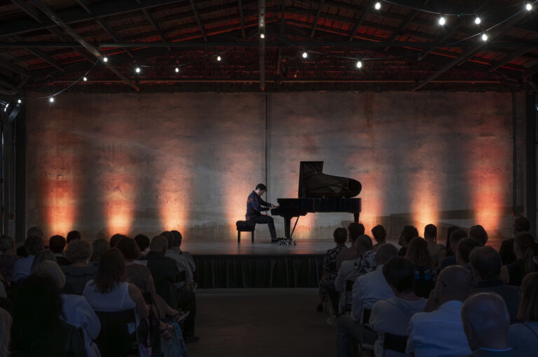 Pianist performing at a grand piano on a dimly lit stage, audience seated in the foreground.
