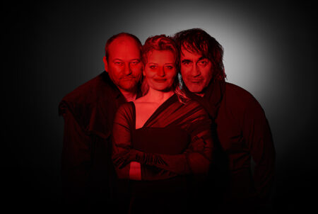 Three people posed together in red-tinted lighting, with a woman at center flanked by two men against a dark backdrop.
