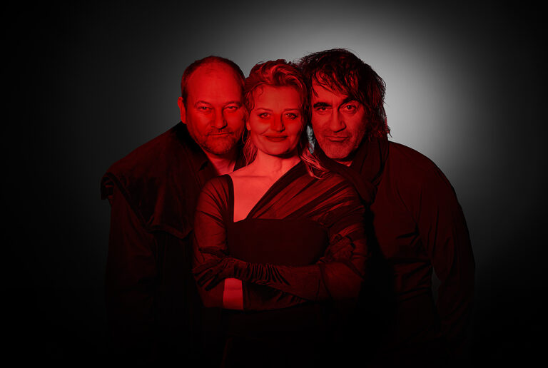 Three people posed together in red-tinted lighting, with a woman at center flanked by two men against a dark backdrop.