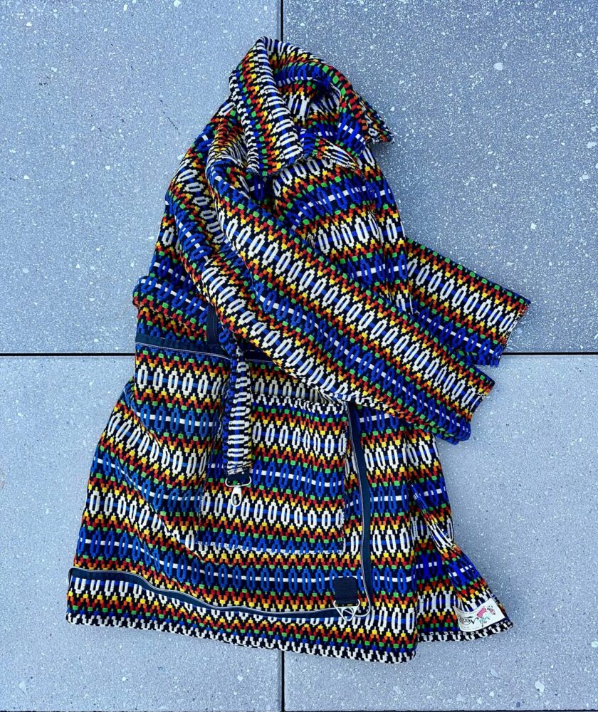 Bright multicolored knitted jacket with geometric stripes laid on a gray pavement, zipper visible and tag at bottom edge