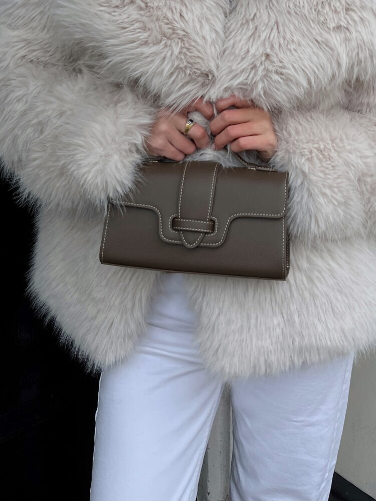 Person in a white fluffy fur coat holding a taupe structured handbag with white stitching, at waist level.