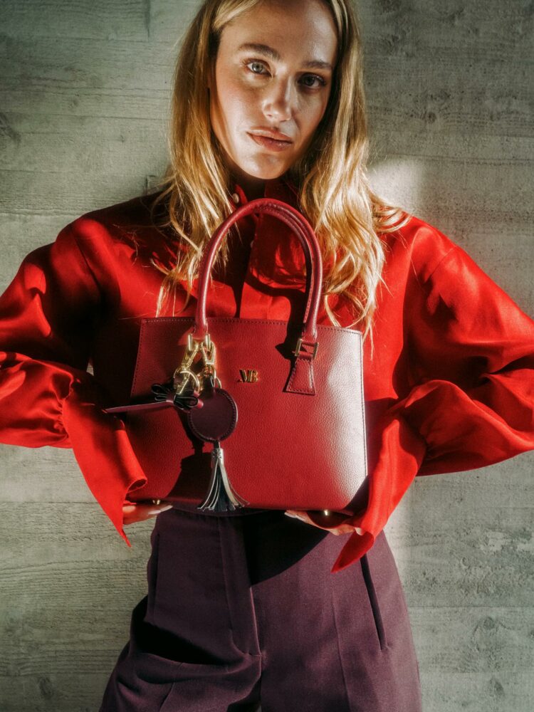 Woman in a red blouse holding a red handbag with gold hardware in front of a concrete wall, facing the camera.