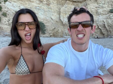 Two friends selfie on a sandy beach in front of rocky cliffs, both making playful faces; woman in a polka dot bikini top, man in a white T-shirt.