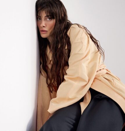Woman with long, wet hair leaning against a white wall in a beige trench and black pants, looking at the camera.