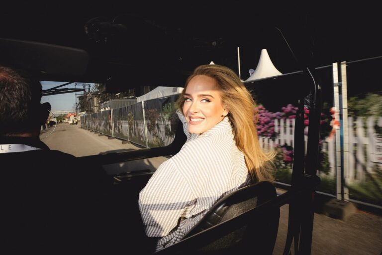Woman with blonde hair wearing a striped shirt sits in a moving vehicle, smiling at the camera as sunlight hits her face.