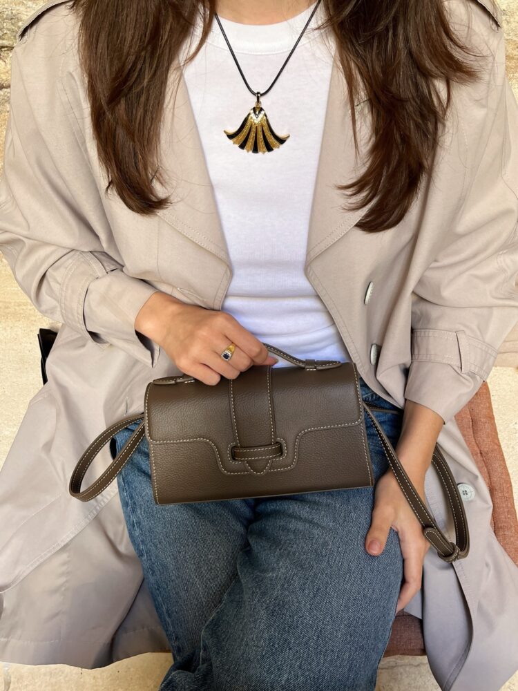 Woman in a beige trench coat over a white top, holding a brown handbag with white stitching; gold fan-shaped necklace visible.