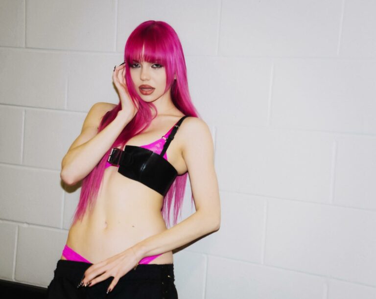 Woman with bright pink hair posing against a white cinder block wall, wearing a black bralette with pink straps and black pants, hand near her face.