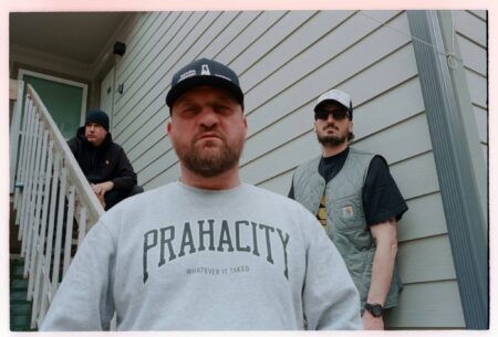 Three men on a residential porch, with one in the foreground wearing a gray sweatshirt and two others behind on the stairs.