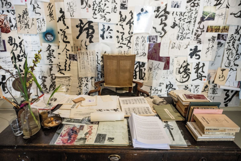 An old wooden desk covered with papers, books, and a glass vase of branches, against a wall full of hanging calligraphy sheets and photos.