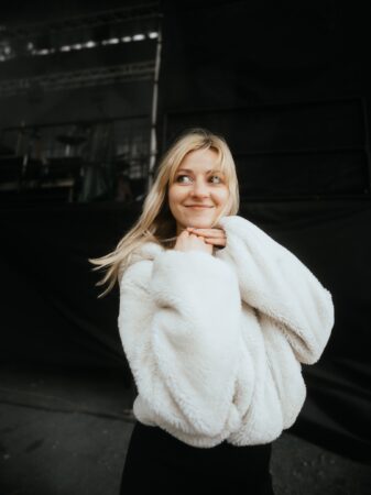 Young woman with blonde hair in a white fluffy coat, smiling and looking upward against a dark stage backdrop, conveying a candid backstage vibe.