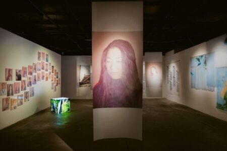Dimly lit art gallery with a tall central panel showing a large portrait of a woman’s face, surrounded by a wall of small square photos to the left.