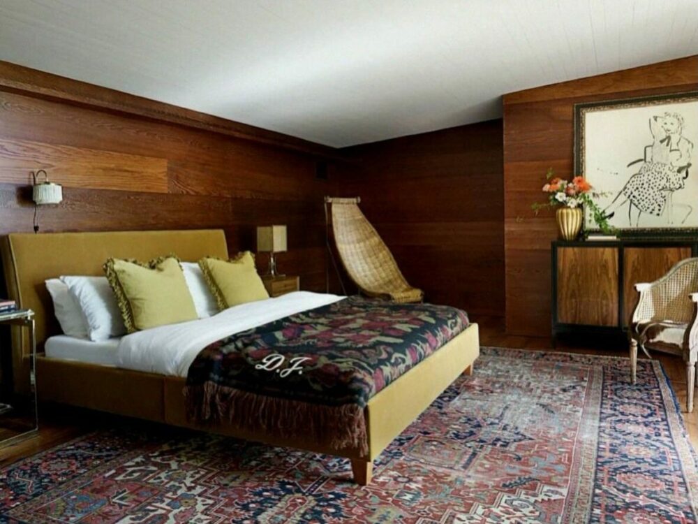A luxurious bedroom with a mustard upholstered bed, floral throw, and Persian rug on a wood-paneled wall. A king bed topped with white bedding and green pillows in a wood-paneled room with a decorative rug and seating.