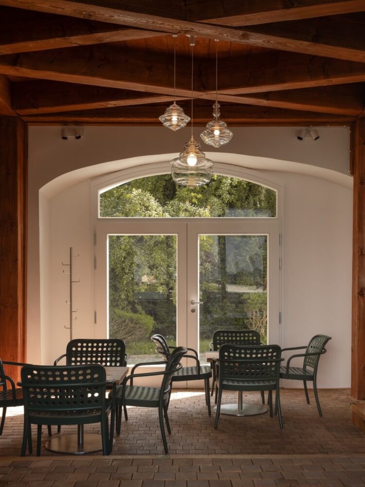 Interior of a cafe with exposed wooden beams, pendant glass lights, and green perforated chairs around round tables by a large arched glass door opening to a garden. You can see trees outside through the door.