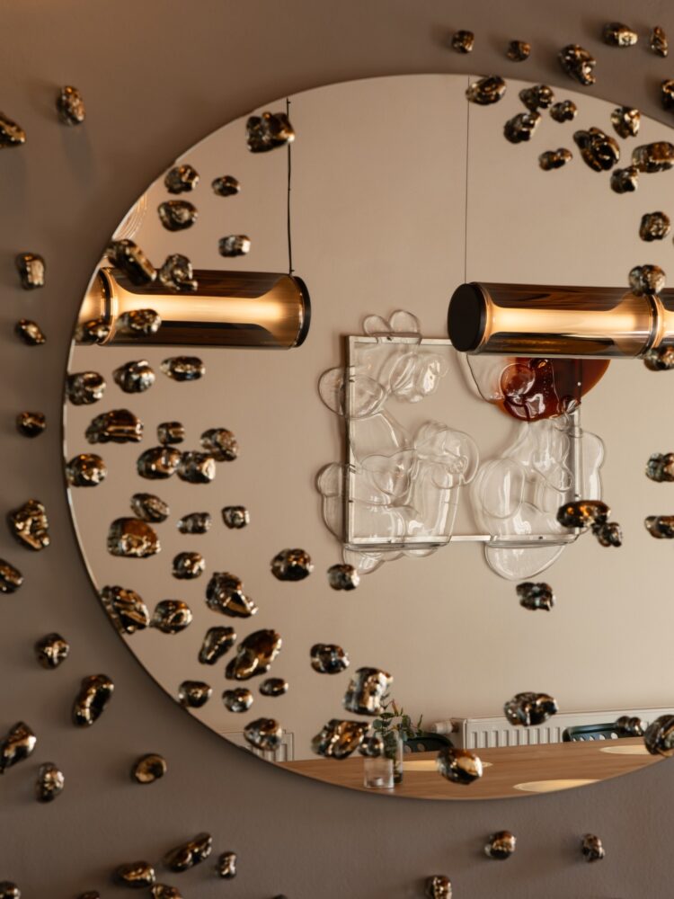 Round wall mirror with warm cylindrical light fixtures and scattered glass orbs; reflection shows a wooden table with a small vase.