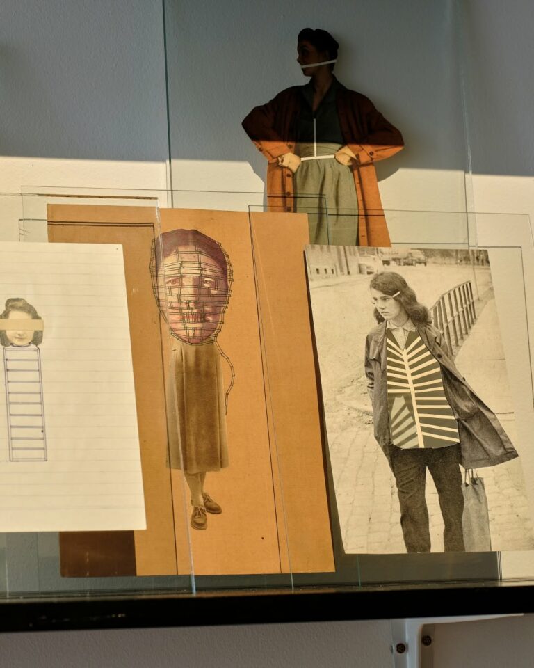 Exhibit display of vintage fashion prints and a life-size cutout wearing an orange coat, arranged behind glass panels.