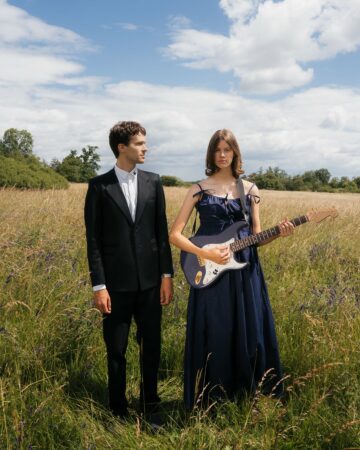 A man in a black suit stands beside a woman in a navy blue dress holding an electric guitar in a sunny, tall‑grass field.