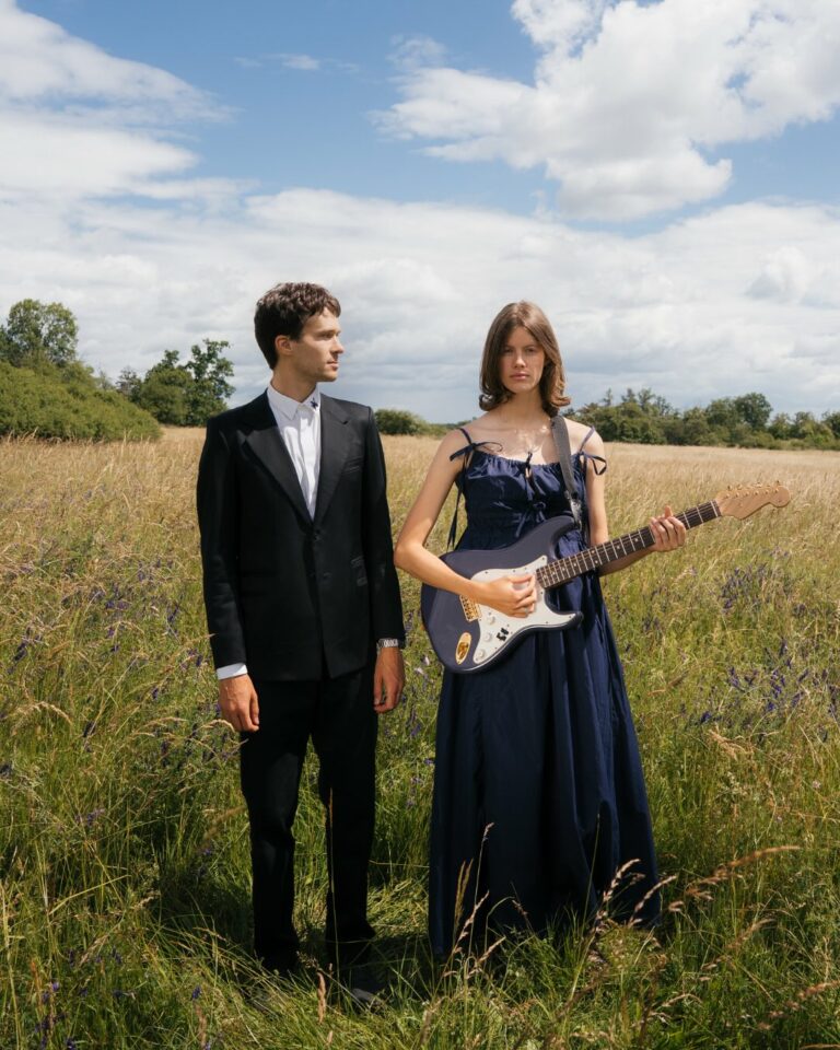 A man in a black suit stands beside a woman in a navy blue dress holding an electric guitar in a sunny, tall‑grass field.