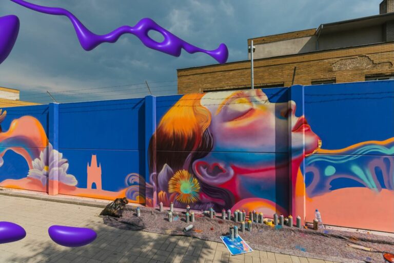 Colorful mural of a woman's face in profile on a blue wall, with spray cans and painting supplies on the ground in front.