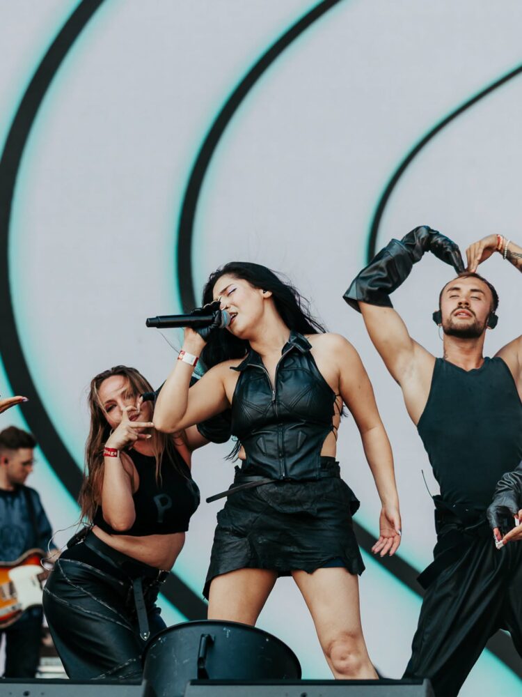Female singer in a black leather outfit belts into a microphone as dancers perform energetically on stage with a curved turquoise backdrop.