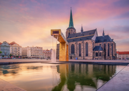 Sunset over a European square with a tall church spire and a gold, rectangular fountain in the foreground reflectively waterlogged pavement prevents glare.