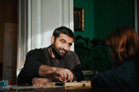 Bearded man smiling as he moves a game piece across a tabletop board while a person with red hair watches.