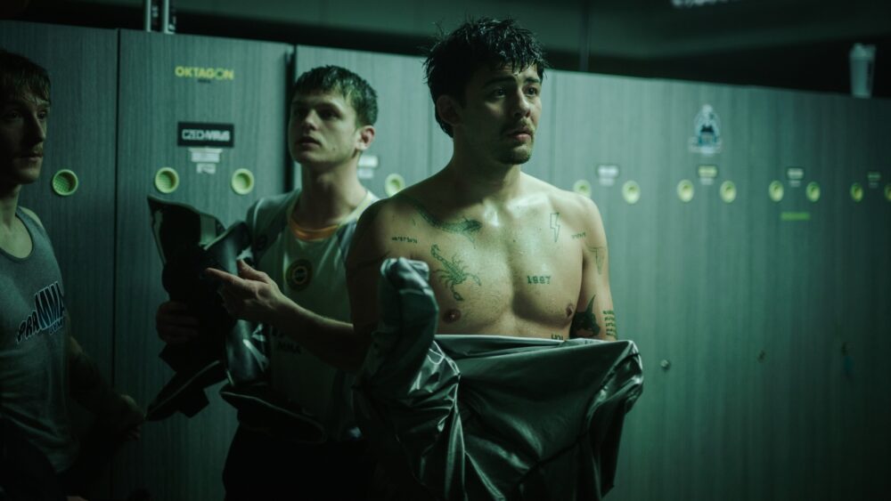 Shirtless man with tattoos in a locker room, adjusting a jacket while two teammates help him.