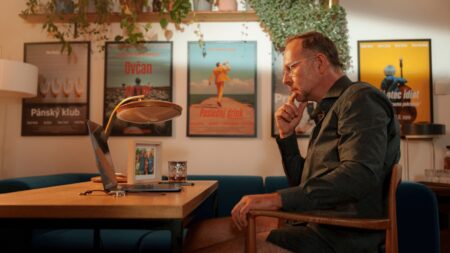Man in glasses sits at a wooden desk with a laptop, hand on chin, in a cozy room with framed posters and plants in the background.