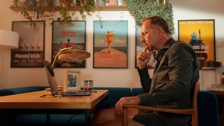 Man in glasses sits at a wooden desk with a laptop, hand on chin, in a cozy room with framed posters and plants in the background.