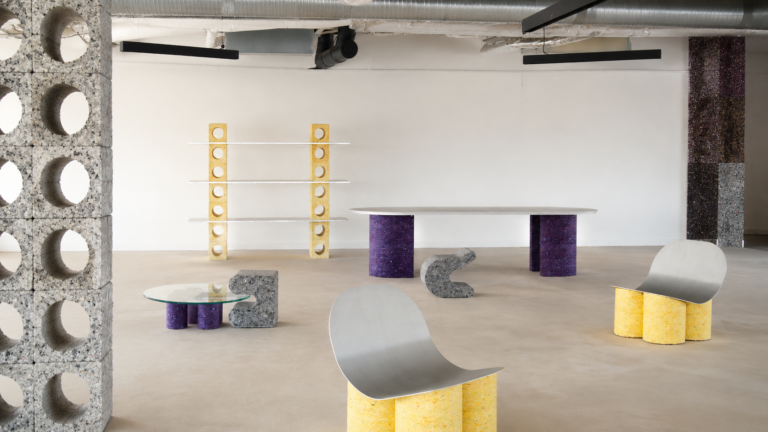 Industrial showroom with modular furniture: yellow cylindrical supports, curved metal seating, glass round table, and tall perforated block walls on left and back.