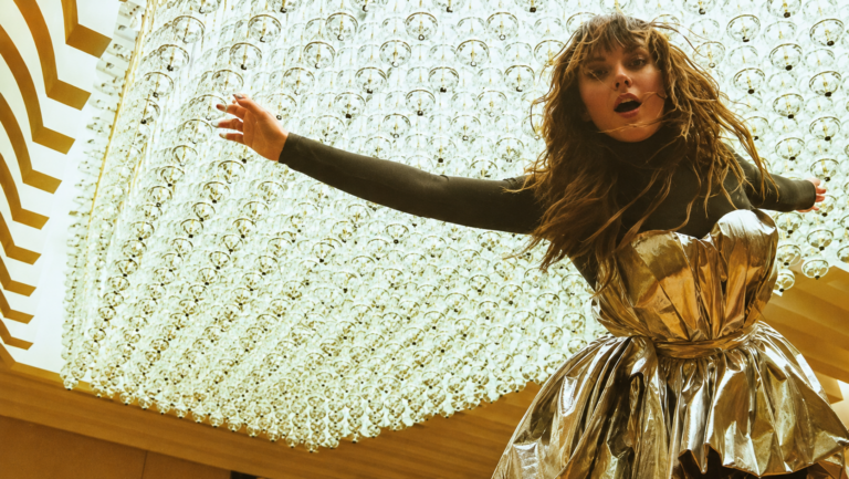 Young woman in a shiny gold dress with arms outstretched, in front of a ceiling installation of hanging glass orbs.