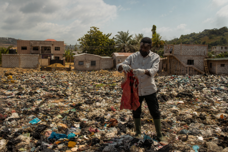 Man wearing gloves and rubber boots sorts through a red shirt in a vast landfill, with unfinished houses in the background.
