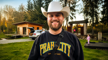 Smiling man wearing a white cowboy hat and black sweatshirt outdoors, with a wooden cabin and kids playing in the background.