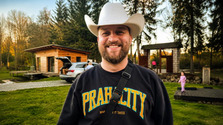 Smiling man wearing a white cowboy hat and black sweatshirt outdoors, with a wooden cabin and kids playing in the background.