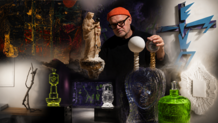 Man in an orange beanie adjusts glass sculptures in a dim art studio with a blue zigzag wall piece in the background.