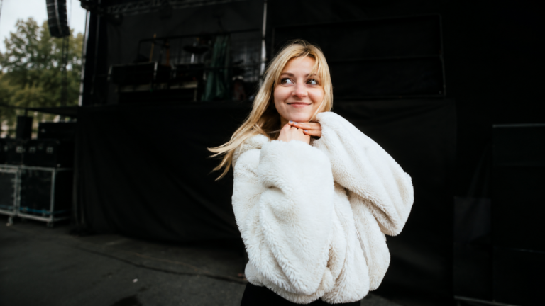 Young woman in a white fluffy jacket posing with her hands under her chin on a backstage stage area.