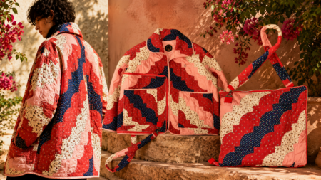Person wearing a pink, red, and navy patchwork quilted jacket stands beside a display of matching quilted bag and tote by pink bougainvillea on stone steps.