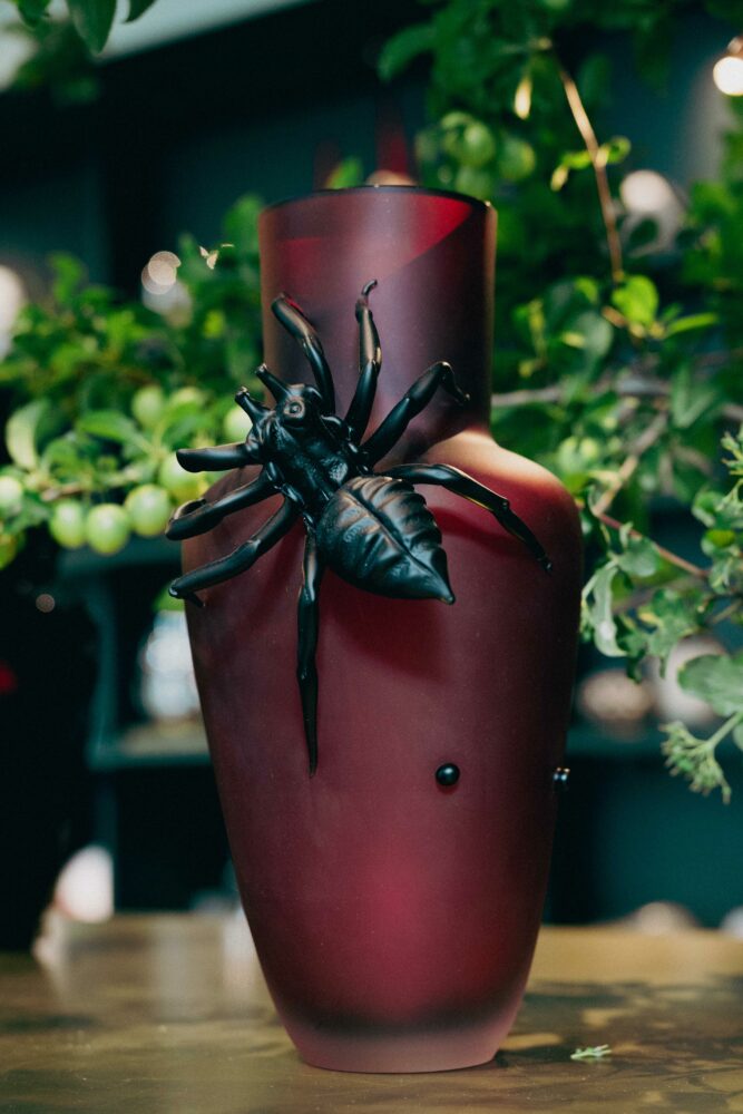 Red ceramic vase with a large black spider ornament attached to its front, greenery in the background.