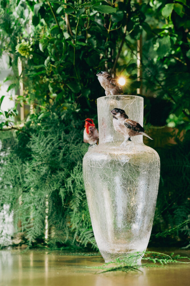 Three small bird sculptures perched on a frosted outdoor vase amid lush green foliage at dusk or night.