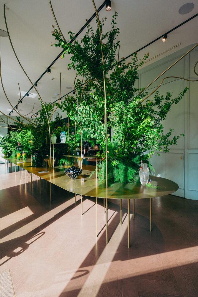 Indoor showroom with dense green plants arranged along a long gold table and curved gold rods overhead, sunlit.