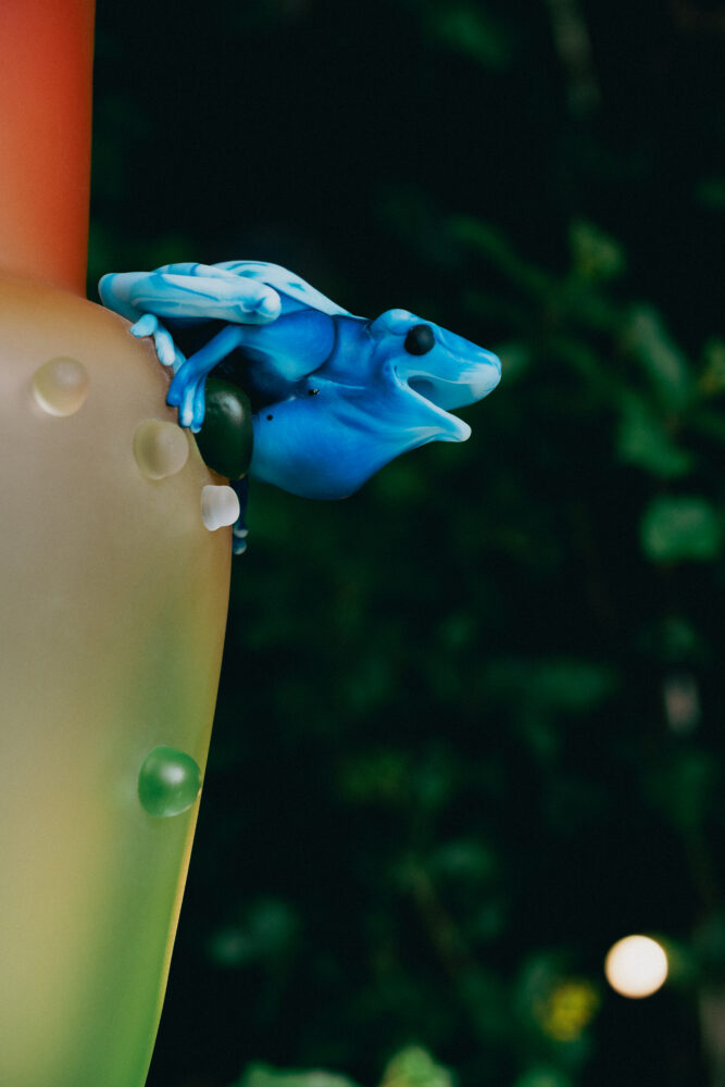 Close-up of a bright blue plastic dragon gripping a beige-green arm adorned with colorful beads against a dark, blurred background.